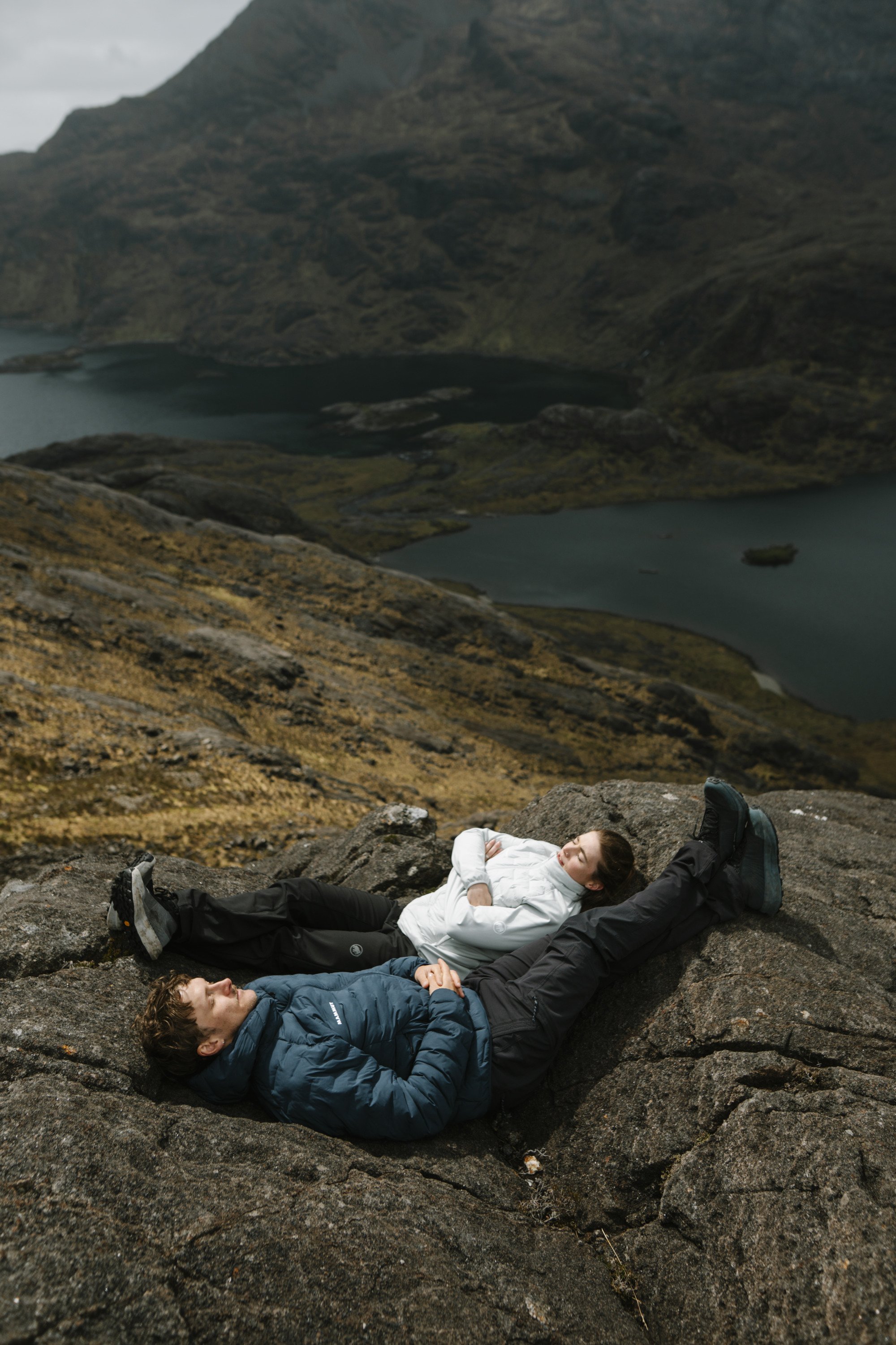 Two people in Mammut outdoor gear relax on lakeside rocks, with scenic mountains in the background, enjoying nature.